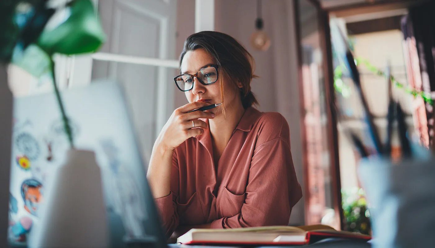 A thoughtful young businesswoman sitting at her desk with an open laptop, reflecting while reading an email and troubleshooting a work issue during long hours.